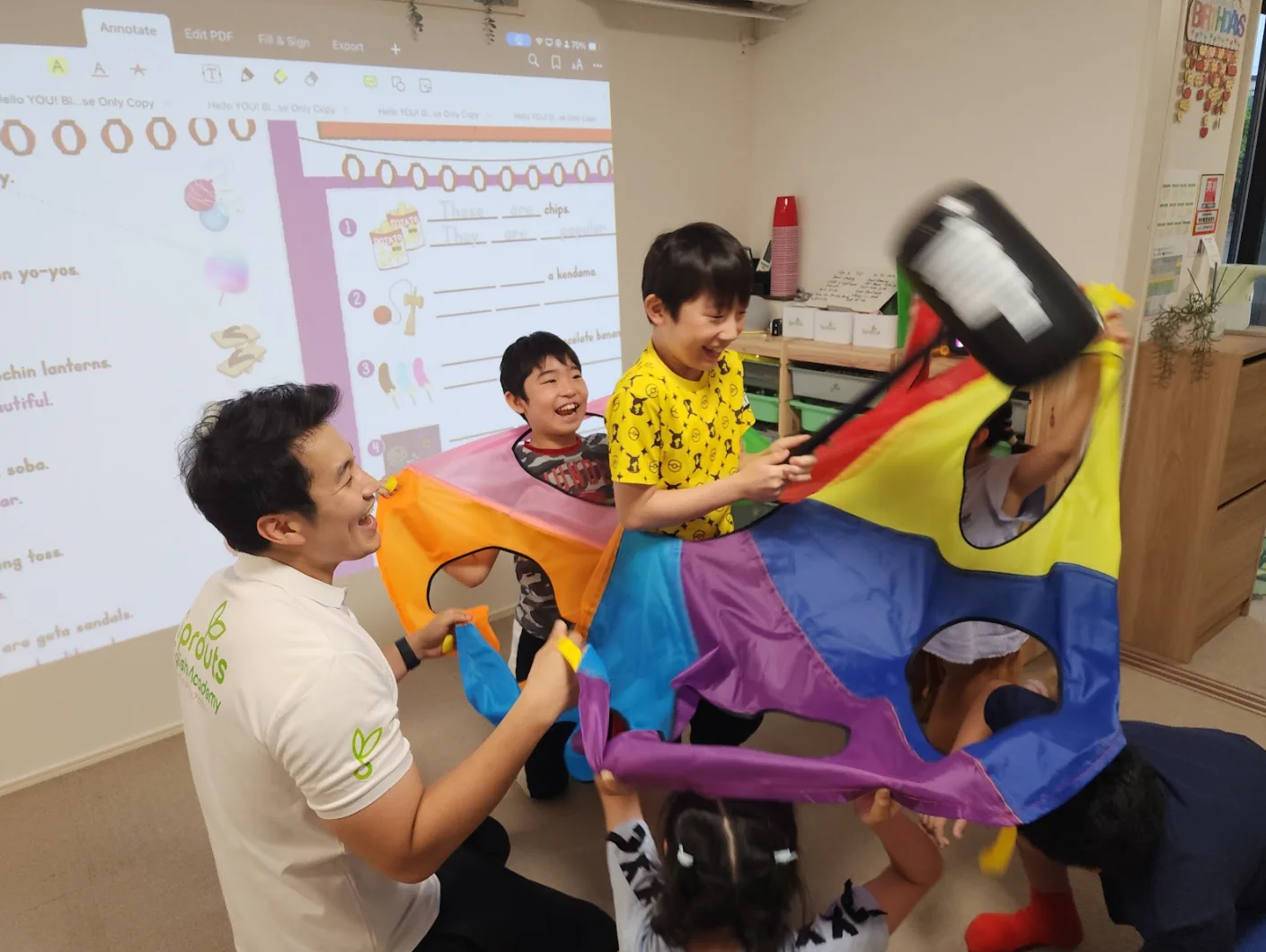 先生と子どもたちがパラシュート遊びのような活動をしながら、英語を楽しく使っている様子